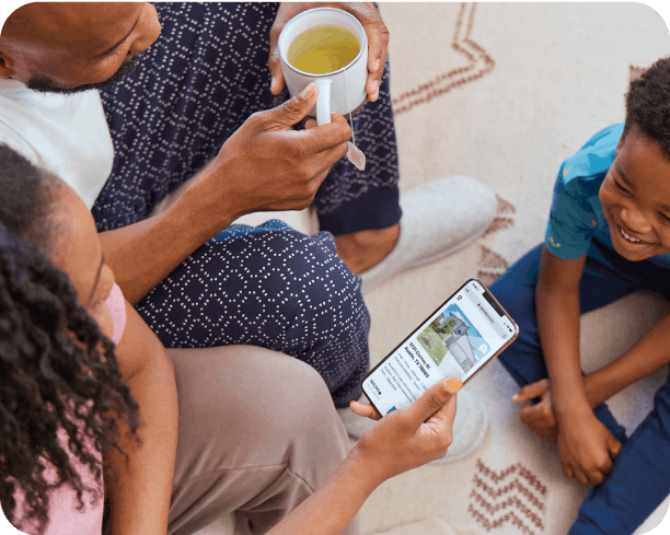 A family relaxing on their rug. Dad holds a cup of tea and mom is on the Opendoor app while their kid smiles up at them.
