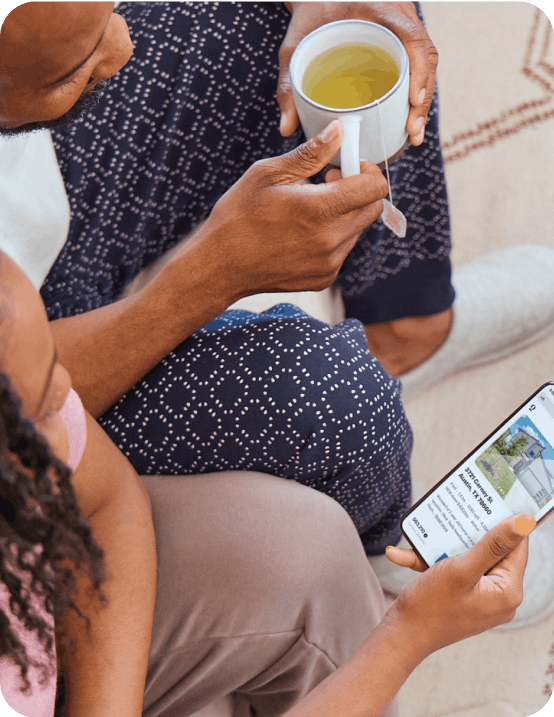 A family relaxing on their rug. Dad holds a cup of tea and mom is on the Opendoor app while their kid smiles up at them.