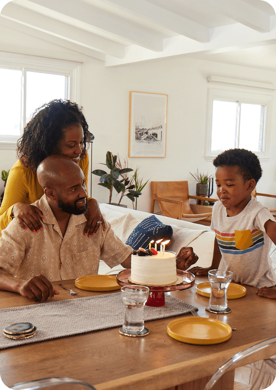 A smiling husband and wife sitting at their living room table with their son as he blows out the candles on his birthday cake.